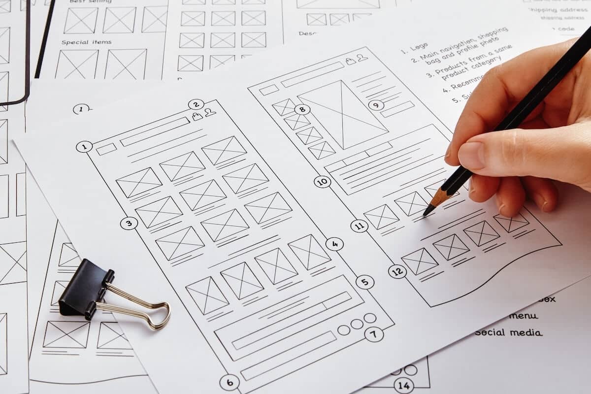 Ein Blatt Papier mit händisch gezeichneten Wireframes. Ein Hand mit Bleistift, die in den Wireframes arbeitet.